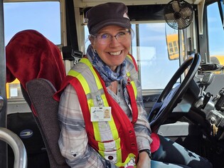 A smiling woman wearing glasses, a grey and red plaid shirt, a high-visibility vest, and a dark-colored cap sits in the driver's seat of a school bus. She has a name tag that reads "Peace River School Division Bus Driver" clipped to her vest. Th