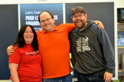 A cheerful group of three adults poses for a photo. The woman, Paul Rowe Jr./Sr. High School Principal Vikki Presakarchuk, is on the left. She has long, dark hair and is wearing a red t-shirt with the words "Teacher Strong" in white script. She