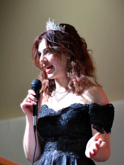 Susie Friesen, a young woman, with shoulder-length auburn hair, wearing a tiara and a black dress, holds a microphone.