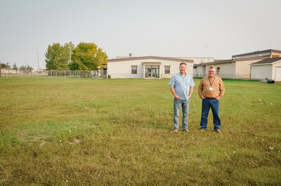 North Peace Commercial Driving Academy Driver Trainers Rick Carter (white male, blue shirt and jeans) and Rick Wearden (white male, brown shirt and jeans) stand in a large grass field