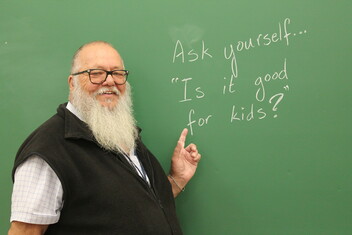 A smiling man with a long white beard and glasses points to a green chalkboard that reads: &quot;Ask yourself... 'Is it good for kids?'&quot;